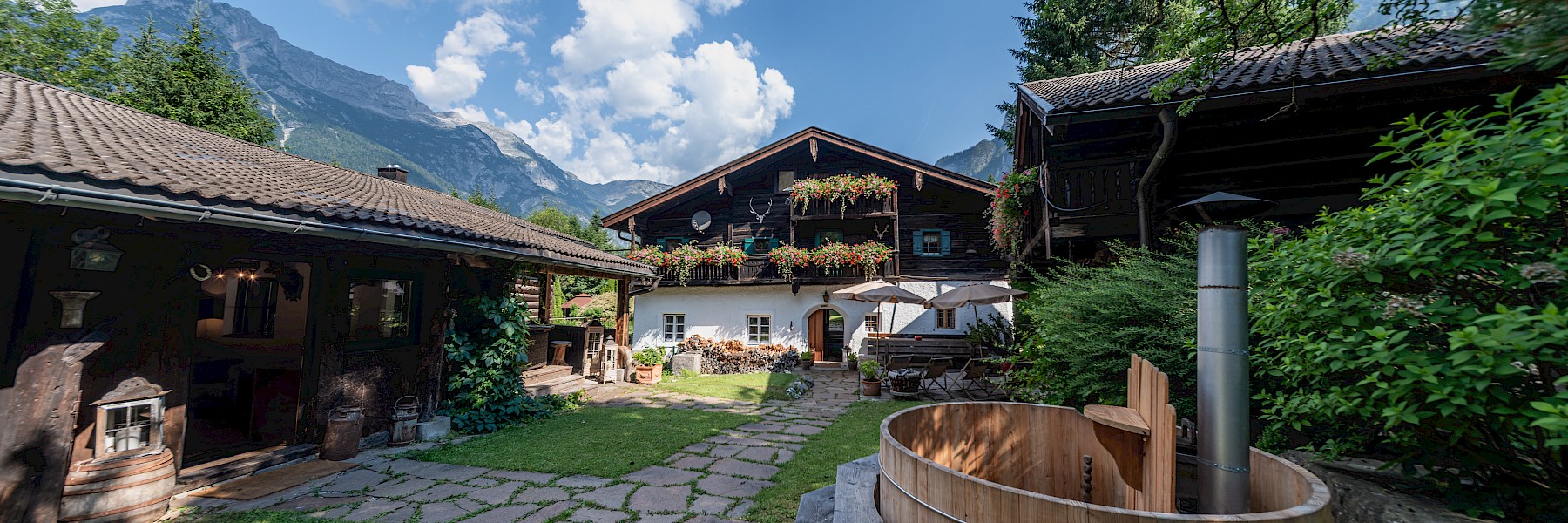 Das Bild zeigt einen idyllischen Innenhof eines traditionellen alpinen Landhauses an einem sonnigen Tag. Im Zentrum steht ein rustikales, dunkles Holzhaus mit üppig blühenden Balkonkästen voller rosa und violetter Blumen. Links und rechts rahmen weitere Gebäude und Pflanzen den Hof ein. Ein Weg aus Natursteinplatten führt über eine gepflegte Rasenfläche zum Hauseingang. Rechts im Bild befindet sich ein runder, hölzerner Badezuber. Im Hintergrund ragen bewaldete Berghänge und schneebedeckte Gipfel in den blauen Himmel. Die Szene wirkt friedlich, gepflegt und typisch für eine ländliche Bergregion.