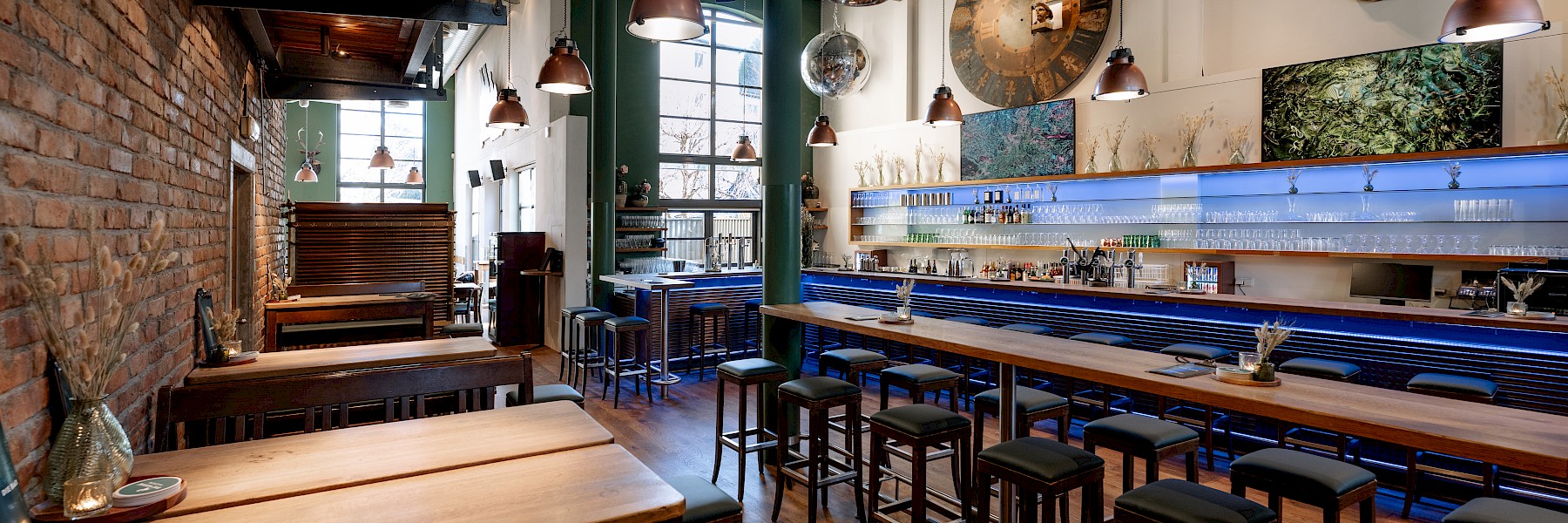 Diese romantische Szene zeigt eine Trauung im Freien, eingebettet in einen schattigen Park mit alten Bäumen. Ein weißer Teppich führt zum Altar, wo das Brautpaar vor einer Traurednerin sitzt. Weiße Stühle für die Gäste sind beidseitig aufgestellt, dekoriert mit zarten Blumenarrangements. Riesige weiße Luftballons, an Schnüren befestigt, verleihen dem Setting eine verspielte Eleganz. Die Atmosphäre wirkt feierlich und zugleich entspannt, mit Gästen in sommerlicher Kleidung, die dem Moment aufmerksam folgen. Im Vordergrund ist eine Frau mit Kinderwagen zu sehen – ein Zeichen dafür, dass hier Generationen zusammenkommen, um die Liebe zu feiern.