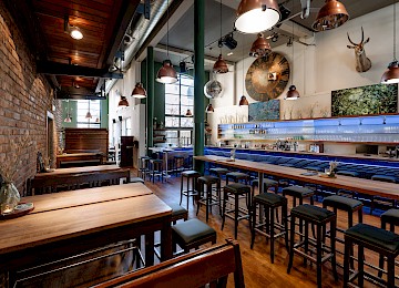 Diese romantische Szene zeigt eine Trauung im Freien, eingebettet in einen schattigen Park mit alten Bäumen. Ein weißer Teppich führt zum Altar, wo das Brautpaar vor einer Traurednerin sitzt. Weiße Stühle für die Gäste sind beidseitig aufgestellt, dekoriert mit zarten Blumenarrangements. Riesige weiße Luftballons, an Schnüren befestigt, verleihen dem Setting eine verspielte Eleganz. Die Atmosphäre wirkt feierlich und zugleich entspannt, mit Gästen in sommerlicher Kleidung, die dem Moment aufmerksam folgen. Im Vordergrund ist eine Frau mit Kinderwagen zu sehen – ein Zeichen dafür, dass hier Generationen zusammenkommen, um die Liebe zu feiern.