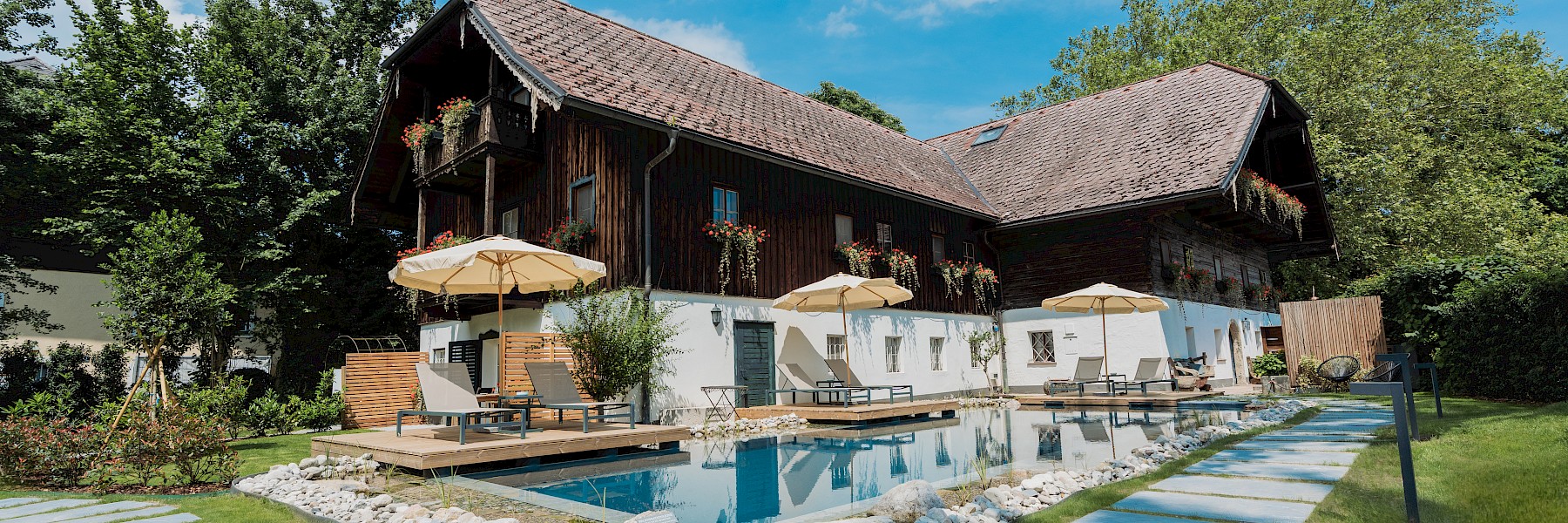 Diese idyllische Gartenszene zeigt ein stimmungsvolles Restaurant im Freien in den Abendstunden. Ein gepflasterter Weg schlängelt sich über eine gepflegte Rasenfläche zu einem liebevoll dekorierten Terrassenbereich, wo Gäste unter großen weißen Sonnenschirmen speisen. Bequeme Liegestühle und Loungemöbel laden zum Verweilen ein, umgeben von blühenden Pflanzen und schattenspendenden Bäumen. Im Hintergrund sorgt ein mit Efeu bewachsenes Haus mit rustikalem Charme für eine romantische Atmosphäre. Warmes Licht und entspannte Gespräche schaffen ein einladendes Ambiente – ideal für ein sommerliches Abendessen oder eine stilvolle Gartenfeier.