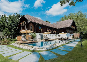 Diese idyllische Gartenszene zeigt ein stimmungsvolles Restaurant im Freien in den Abendstunden. Ein gepflasterter Weg schlängelt sich über eine gepflegte Rasenfläche zu einem liebevoll dekorierten Terrassenbereich, wo Gäste unter großen weißen Sonnenschirmen speisen. Bequeme Liegestühle und Loungemöbel laden zum Verweilen ein, umgeben von blühenden Pflanzen und schattenspendenden Bäumen. Im Hintergrund sorgt ein mit Efeu bewachsenes Haus mit rustikalem Charme für eine romantische Atmosphäre. Warmes Licht und entspannte Gespräche schaffen ein einladendes Ambiente – ideal für ein sommerliches Abendessen oder eine stilvolle Gartenfeier.