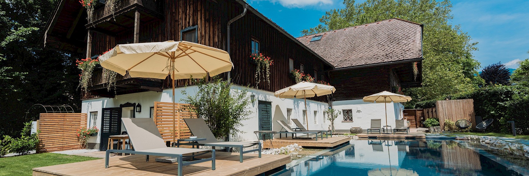 Diese idyllische Gartenszene zeigt ein stimmungsvolles Restaurant im Freien in den Abendstunden. Ein gepflasterter Weg schlängelt sich über eine gepflegte Rasenfläche zu einem liebevoll dekorierten Terrassenbereich, wo Gäste unter großen weißen Sonnenschirmen speisen. Bequeme Liegestühle und Loungemöbel laden zum Verweilen ein, umgeben von blühenden Pflanzen und schattenspendenden Bäumen. Im Hintergrund sorgt ein mit Efeu bewachsenes Haus mit rustikalem Charme für eine romantische Atmosphäre. Warmes Licht und entspannte Gespräche schaffen ein einladendes Ambiente – ideal für ein sommerliches Abendessen oder eine stilvolle Gartenfeier.
