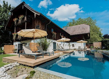 Diese idyllische Gartenszene zeigt ein stimmungsvolles Restaurant im Freien in den Abendstunden. Ein gepflasterter Weg schlängelt sich über eine gepflegte Rasenfläche zu einem liebevoll dekorierten Terrassenbereich, wo Gäste unter großen weißen Sonnenschirmen speisen. Bequeme Liegestühle und Loungemöbel laden zum Verweilen ein, umgeben von blühenden Pflanzen und schattenspendenden Bäumen. Im Hintergrund sorgt ein mit Efeu bewachsenes Haus mit rustikalem Charme für eine romantische Atmosphäre. Warmes Licht und entspannte Gespräche schaffen ein einladendes Ambiente – ideal für ein sommerliches Abendessen oder eine stilvolle Gartenfeier.