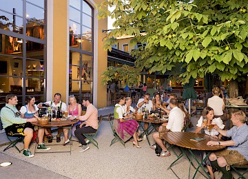 Die Aufnahme zeigt das elegante Eingangsschild des „Rosewood Munich“, montiert an einer Fassade aus rustikalem Sandstein. Die goldenen, stilvoll in das dunkle Metallschild eingelassenen Buchstaben heben sich kontrastreich von der hellen Natursteinwand ab und unterstreichen den luxuriösen Charakter des Gebäudes. Die Architektur kombiniert klassische Elemente mit moderner Ästhetik, was dem Hotel einen exklusiven und zugleich einladenden Auftritt verleiht. Im Vordergrund sind gepflegte Pflanzgefäße zu erkennen, die den gehobenen Stil zusätzlich unterstreichen. Das Bild vermittelt einen ersten Eindruck von Eleganz, Klasse und hochwertigem Design mitten im Herzen Münchens.