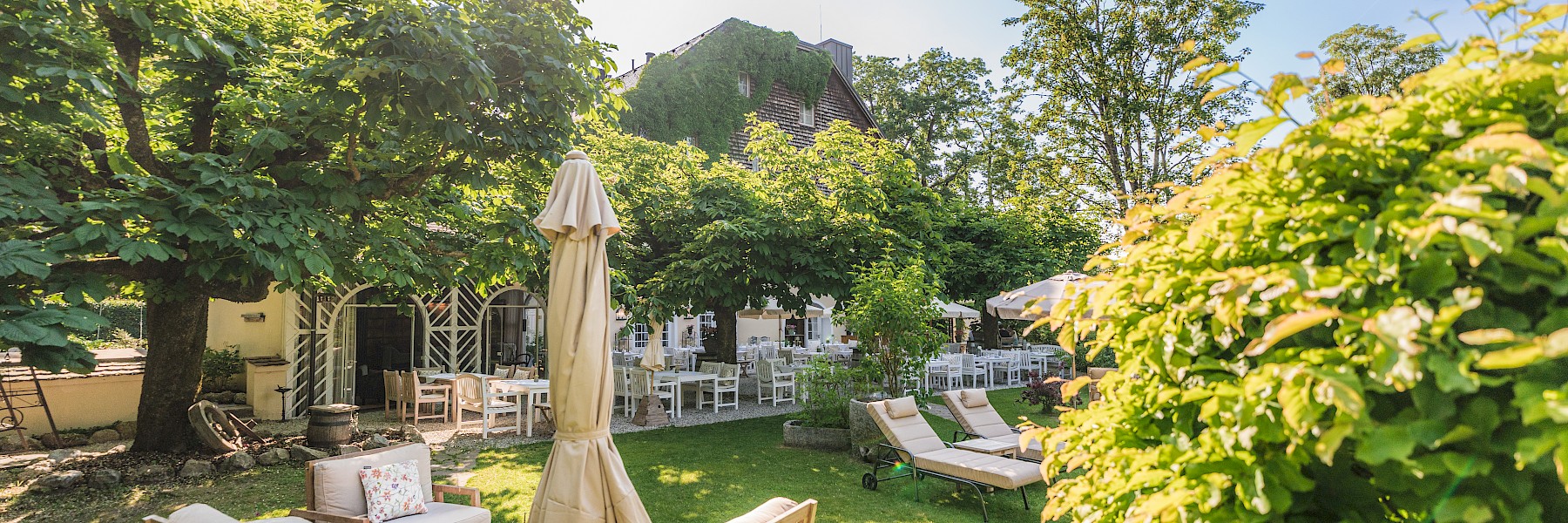 Gastgarten Schlosswirt zu Anif (c) Gassner Gastronomie, Foto Hechtfisch: Der idyllische Gastgarten lädt zum Entspannen und Genießen ein. Bequeme Sitzmöbel auf der Terrasse, Liegen auf der gepflegten Rasenfläche und zahlreiche Plätze im schattigen Gastgarten bieten Erholung inmitten von Bäumen und üppigem Grün. Das charmante Gebäude im Hintergrund rundet die ruhige und einladende Atmosphäre ab.