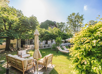 Gastgarten Schlosswirt zu Anif (c) Gassner Gastronomie, Foto Hechtfisch: Der idyllische Gastgarten lädt zum Entspannen und Genießen ein. Bequeme Sitzmöbel auf der Terrasse, Liegen auf der gepflegten Rasenfläche und zahlreiche Plätze im schattigen Gastgarten bieten Erholung inmitten von Bäumen und üppigem Grün. Das charmante Gebäude im Hintergrund rundet die ruhige und einladende Atmosphäre ab.