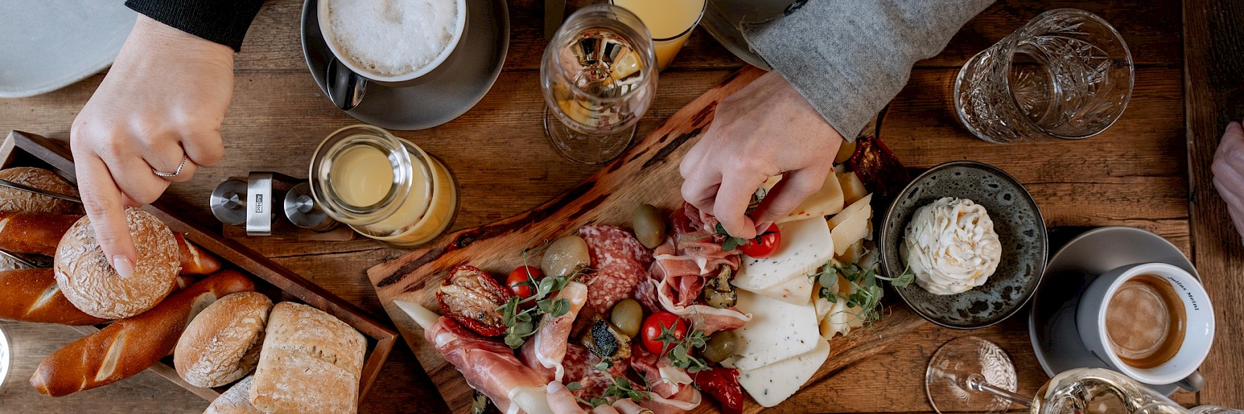 Brunch @ Gassner Gastronomie, Foto Hechtfisch: Ein liebevoll gedeckter Holztisch lädt zu einem genussvollen Frühstück oder Brunch ein. Frische Brötchen, Brezeln und Brot stehen bereit, dazu eine reichhaltige Platte mit feinem Käse, Schinken, Salami, Oliven und frischen Tomaten. Ergänzt wird das Angebot durch belegte Brote, Kaffee mit Milchschaum, Espresso, Orangensaft und weitere Getränke. Mehrere Hände greifen gemeinsam nach den Köstlichkeiten – ein Zeichen für geselliges Beisammensein und entspannten Genuss. Die warme Atmosphäre und die hochwertige Auswahl machen Lust, gemeinsam Zeit zu verbringen und den Tag genussvoll zu beginnen.