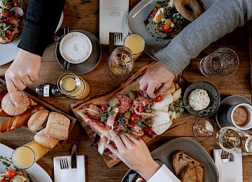 Brunch @ Gassner Gastronomie, Foto Hechtfisch: Ein liebevoll gedeckter Holztisch lädt zu einem genussvollen Frühstück oder Brunch ein. Frische Brötchen, Brezeln und Brot stehen bereit, dazu eine reichhaltige Platte mit feinem Käse, Schinken, Salami, Oliven und frischen Tomaten. Ergänzt wird das Angebot durch belegte Brote, Kaffee mit Milchschaum, Espresso, Orangensaft und weitere Getränke. Mehrere Hände greifen gemeinsam nach den Köstlichkeiten – ein Zeichen für geselliges Beisammensein und entspannten Genuss. Die warme Atmosphäre und die hochwertige Auswahl machen Lust, gemeinsam Zeit zu verbringen und den Tag genussvoll zu beginnen.