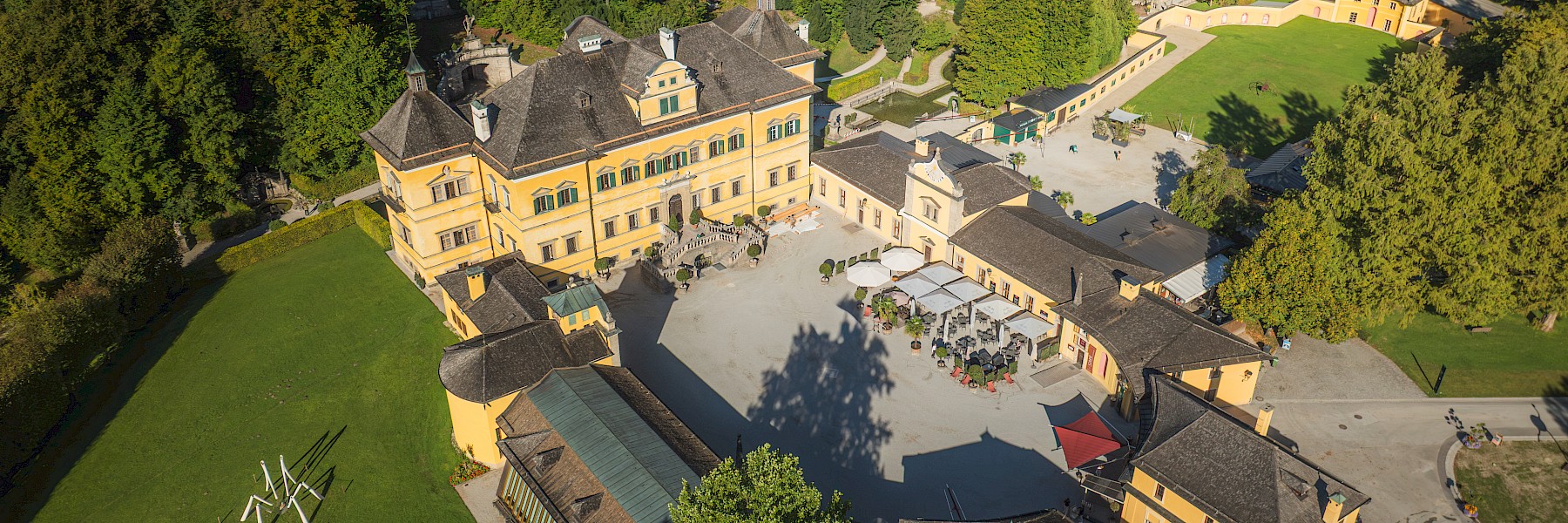 Luftaufnahme Schloss Hellbrunn @ Gassner Gastronomie, Foto Hechtfisch: Das Bild zeigt eine Drohnenaufnahme von Schloss Hellbrunn mit seiner weitläufigen Schlossanlage. Das barocke Schloss mit seiner gelben Fassade liegt eingebettet in eine große Parklandschaft mit alten Bäumen, gepflegten Wiesen und Spazierwegen. Im Innenhof befinden sich Sitzplätze für Gäste. Rund um das Schloss erstrecken sich Felder und Wälder, die eine ruhige, naturnahe Umgebung schaffen. Die Aufnahme vermittelt einen eindrucksvollen Überblick über das historische Anwesen und seine großzügigen Außenanlagen.