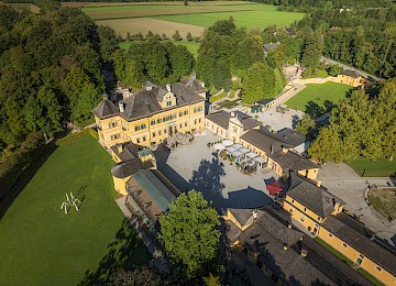 Luftaufnahme Schloss Hellbrunn @ Gassner Gastronomie, Foto Hechtfisch: Das Bild zeigt eine Drohnenaufnahme von Schloss Hellbrunn mit seiner weitläufigen Schlossanlage. Das barocke Schloss mit seiner gelben Fassade liegt eingebettet in eine große Parklandschaft mit alten Bäumen, gepflegten Wiesen und Spazierwegen. Im Innenhof befinden sich Sitzplätze für Gäste. Rund um das Schloss erstrecken sich Felder und Wälder, die eine ruhige, naturnahe Umgebung schaffen. Die Aufnahme vermittelt einen eindrucksvollen Überblick über das historische Anwesen und seine großzügigen Außenanlagen.